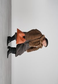 Person som sitter på huk, iklädd en brun pälsjacka, mörka byxor, svarta stövlar och håller en orange handväska mot en enkel grå bakgrund.