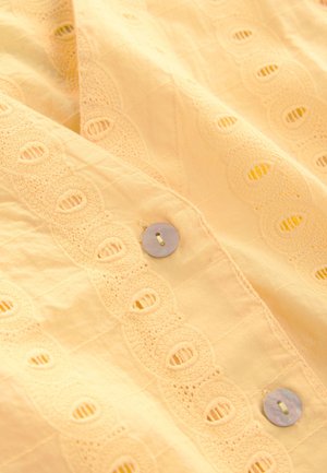 Puha sárga pamutszövet hímzett, hullámos mintával. Az elején kör alakú anyajegyre emlékeztető gombok találhatók.