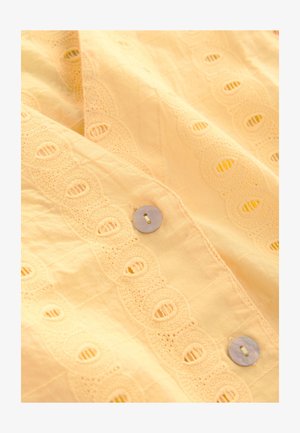 Puha sárga pamutszövet hímzett, hullámos mintával. Az elején kör alakú anyajegyre emlékeztető gombok találhatók.