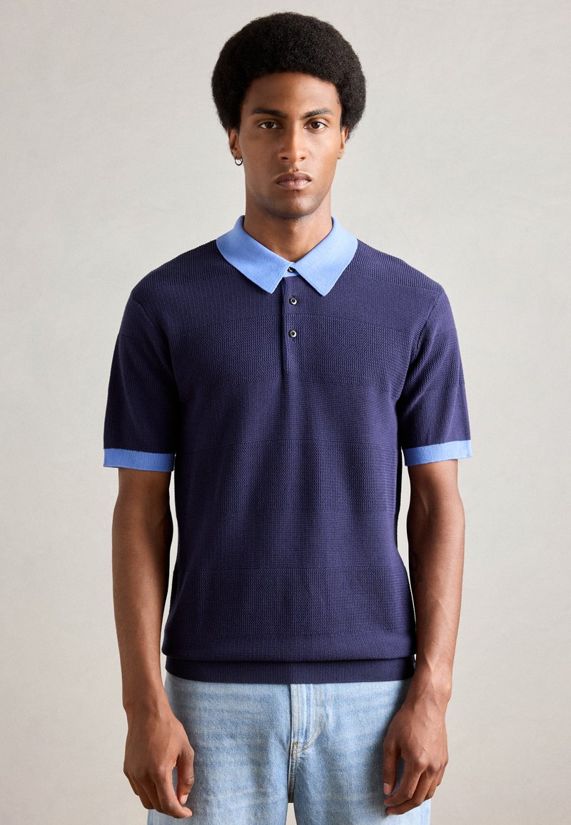 Marinblå kortärmad polo med ljusblå krage och muddar, textil med textur som har horisontella ränder, och treknäppt krage.