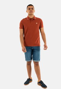 Polo shirt color ruggine con colletto a costine e piccolo logo, abbinato a pantaloni corti di jeans e sneakers scure con dettagli arancioni.