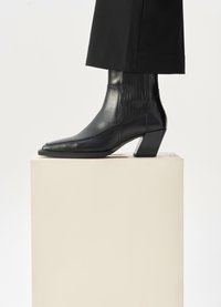 Μαύρη δερμάτινη μπότες κάτω από τον αστράγαλο με μυτερή μύτη, πλαϊνά ελαστικά πάνελ και χοντρό τακούνι, τοποθετημένη σε έναν ανοιχτό πεζόβραχο μπροστά σε μια ανοιχτόχρωμη φόντο.