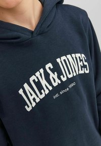 Sudadera azul marino con un logo blanco de "JACK & JONES" y el texto "intl. desde 1990". Fabricada con un tejido suave, cuenta con un bolsillo tipo canguro y capucha.