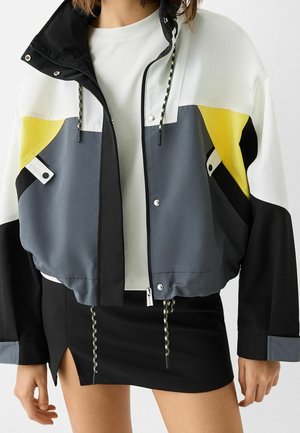 Vrouw draagt een colorblock-jas in wit, geel, grijs en zwart over een wit topje en een zwarte rok met een zijsplit en trekkoorddetails.