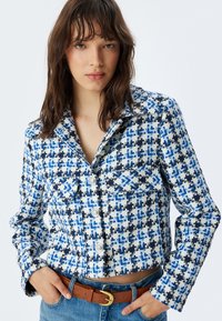 Korta jacka i ett strukturerat blått och vitt hundtandsmönster, med silverknappar och två bröstfickor, kombinerat med ett brunt bälte.