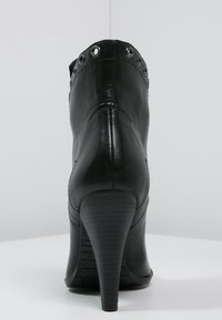 Černá kožená kotníková bota na vysokém podpatku s kovovými oky kolem horního okraje, zobrazená ze zadní strany na bílé ploše.