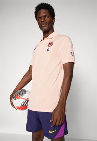 Male soccer player holding white and black ball, wearing light pink polo with club logo and navy shorts with yellow and purple accents.