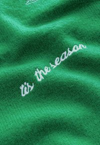 Žalios medžiagos audinys pasižymi tekstūrinio audimo stiliumi su balta siuvinėta užrašu "laikas šventėms". Medžiaga atrodo minkšta ir jauki.