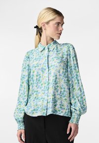 Donna con capelli biondi raccolti da un nastro nero, che indossa una camicetta a maniche lunghe a sbuffo di colore azzurro chiaro, verde e bianco, abbinata a pantaloni neri.