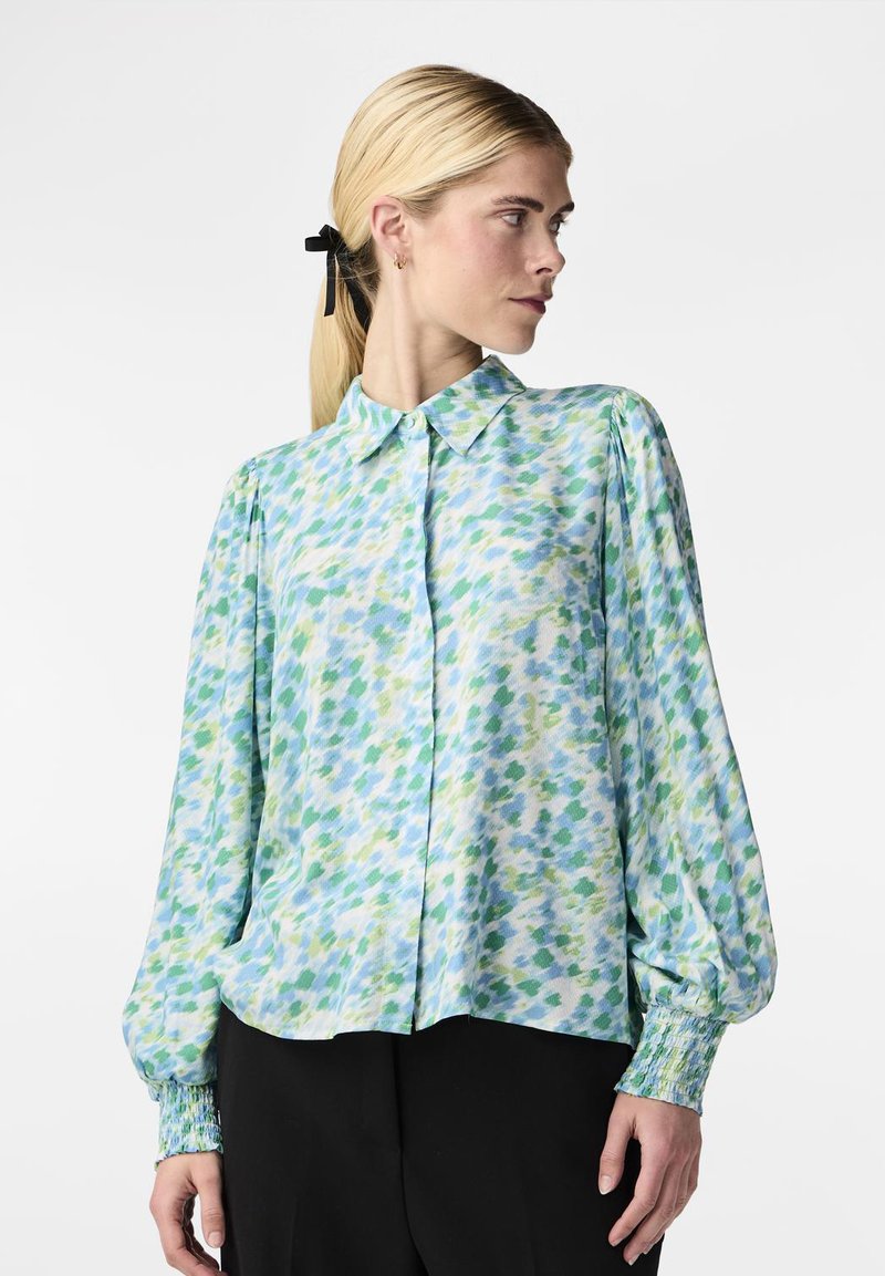 Donna con capelli biondi raccolti da un nastro nero, che indossa una camicetta a maniche lunghe a sbuffo di colore azzurro chiaro, verde e bianco, abbinata a pantaloni neri.