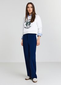 Felpa bianca corta con scritte verdi, abbinata a pantaloni navy a gamba larga. Sneakers bianche completano il look, mostrando uno stile sportivo.