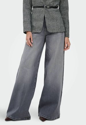 Jean en denim gris délavé à jambes larges, porté avec un blazer gris ceinturé, des chaussures pointues bordeaux et des bagues argentées aux deux mains.