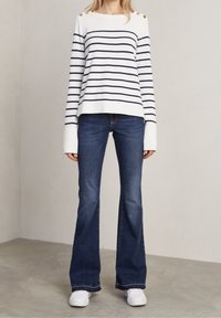 Randig vit och marinblå tröja med guldknappdetaljer på axlarna, i kombination med mörkblå utsvängda jeans och vita sneakers.