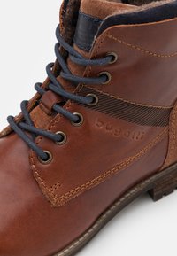 Brązowe skórzane buty za kostkę ze sznurowaniem, z metalowymi oczkami, czarnymi sznurowadłami, wyściełanym kołnierzem i wytłoczonym logo "bugatti" na bocznej ściance.