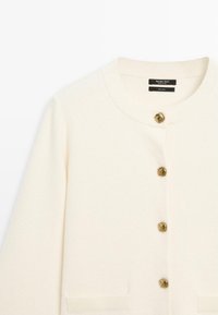 Crème wollen cardigan met een ronde halslijn, vijf metalen knopen met een geometrisch ontwerp en twee voorzakken.