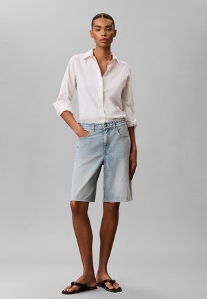 Kvinde stående med den ene hånd i lommen iført en hvid skjorte med knapper, lyseblå denim shorts til knæet og sorte flip-flops.