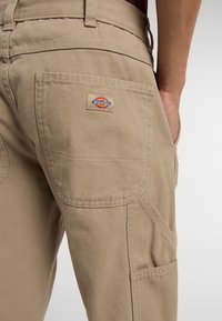 Béžové pracovní kalhoty vyrobené z odolné tkaniny, s několika kapsami a logem na zadní straně. Design zahrnuje zesílené švy.