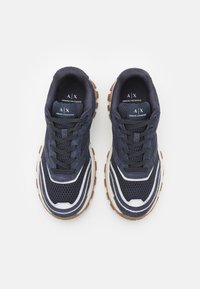 Chaussures de sport navy avec tige en mesh, accents blancs et crème, bout arrondi et semelle en caoutchouc marron avec grip texturé.