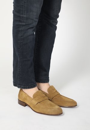 Tan suède instappers met een gladde textuur, voorzien van een decoratieve pennyband en een lage houten hak, gecombineerd met donkerdenim jeans.
