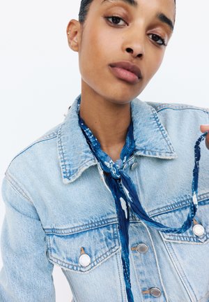 Light blue denim jacket with silver buttons and two chest pockets, paired with a blue patterned scarf tied around the neck.