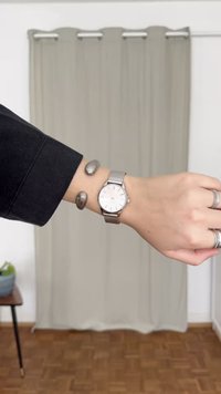 Srebrny zegarek na siateczkowym pasku z białą tarczą i smukłymi znacznikami godzin, noszony na nadgarstku obok dwóch gładkich metalowych bransoletek.