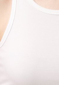 Top de tirantes blanco con textura acanalada y escote redondeado, confeccionado en una suave y elástica tela para mayor comodidad. La vista de cerca resalta la textura y los detalles de las costuras.
