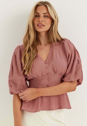 Jeune femme aux longs cheveux blonds portant un chemisier mauve avec des manches bouffantes et des boutons recouverts de tissu, debout devant un fond uni.