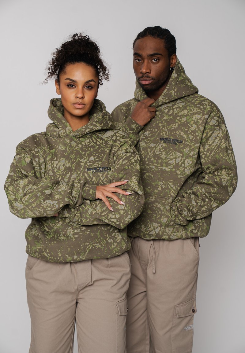 Les sweats à capuche vert olive présentent un motif camouflage feuillu texturé, avec une poche kangourou et des cordons de serrage sur la capuche, associés à un pantalon cargo beige.