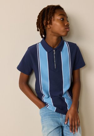 Ung person med korte dreadlocks iført en mørkeblå polo med lyseblå lodrette striber og lyseblå jeans, hænderne i lommerne.