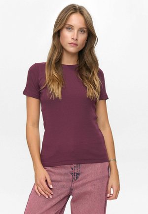 Ung kvinna med långt vågigt hår, iförd en figurnära lila kortärmad topp och högmidjade rosa jeans, stående mot en vit bakgrund.