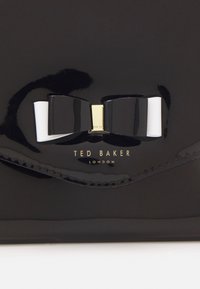 Černá peněženka z lakované kůže s lesklým povrchem. Obsahuje černou a bílou mašličku jako ozdobu a zlatý detail loga, se švem podél okrajů.