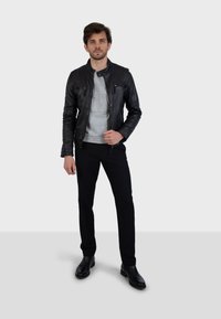 Mladý muž stojí čelom vpred, oblečený v čiernej koženej bunde, šedom tričku, čiernych nohaviciach a čiernych boots na pozadí s jednoduchým pozadím.