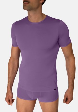 Mann trägt eng anliegendes, lila geripptes Kurzarmshirt und passende Boxershorts und steht vor einem schlichten Hintergrund.