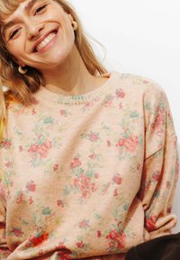 Pull rose clair avec un motif floral aux nuances de rouge et de vert. Texture douce et côtelée avec un col rond. Coupe ample et confortable.