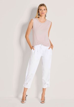 Frau steht vor einer beigen Wand und trägt ein ärmelloses, hellrosa Oberteil, weiße, verkürzte Hose mit geknöpften Manschetten, silberne High-Heel-Sandalen und auffällige Ohrringe.