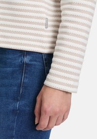 Gestreiftes Langarmshirt in Weiß und Beige, aus strukturiertem Stoff gefertigt. Sichtbares Etikett an der Seite, kombiniert mit blauer Jeans.