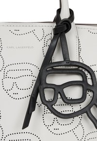 Valge nahast käekott, millel on aukudega näo kujundused, sisaldab musta kummist prillide võlu. Käekotil on graveeritud "Karl Lagerfeld".