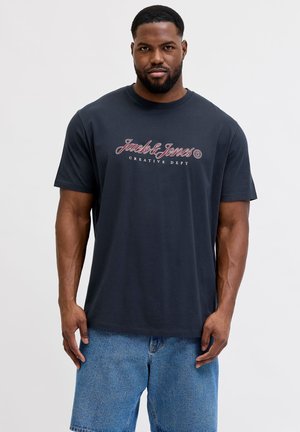 Man met kort haar en baard, die een zwarte "Jack & Jones Creative Dept" t-shirt en blauwe denim short draagt, staand tegen een effen achtergrond.