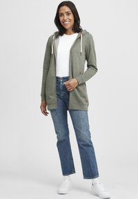 Grønn lang hettejakke laget av mykt stoff, med glidelås foran, snorhette og subtile brune detaljer. Kombinert med blå jeans og hvite joggesko.