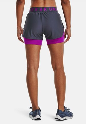 Persona que lleva pantalones cortos deportivos en capas de color gris oscuro y púrpura, y zapatos de correr azul marino, de pie de espaldas sobre un fondo blanco.