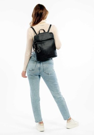 Vrouw met een beige trui, lichtblauwe jeans, witte sneakers en een zwarte leren rugzak, staand met haar rug naar de camera.