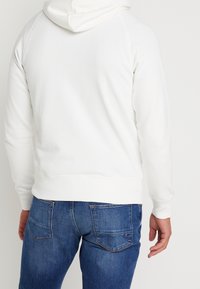 Felpa bianca con cappuccio dal tessuto liscio e orlo a coste, abbinata a jeans in denim blu con tasche posteriori e leggera scoloritura.