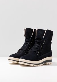 Botas de tobillo de ante azul marino oscuro con punteras redondeadas, parte delantera con cordones, forro acolchado y suelas de goma crema contrastantes con textura.