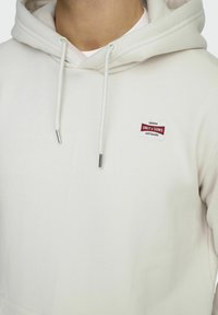 Helle beige Kapuzenjacke mit Kordeln und einem kleinen roten und weißen Etikett mit dem Schriftzug "ONLY & SONS". Weicher Stoff mit lässiger Passform.