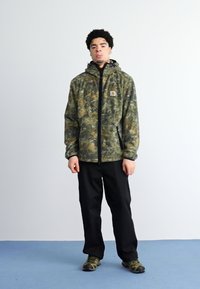 Jachetă de camuflaj cu glugă, fabricată dintr-un material ușor, cu fermoar frontal și buzunare laterale. Purtați cu pantaloni negri și încălțăminte.