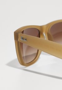 Ochelari de soare Ray-Ban cu un cadru mat de culoare maronie, lentile maronii cu degrade și accent logo pe templu. Design unghiular cu brațe groase.