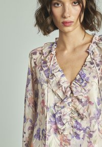 Blumenbluse aus leichtem Stoff mit Rüschenausschnitt, pastellfarbenen Tönen und dekorativen Knöpfen, die ein durchgehendes Blumenmuster zeigt.