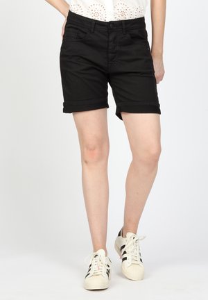 Schwarze Baumwollshorts mit umgeschlagenen Säumen, mit Fronttaschen und Knopfverschluss. Kombiniert mit weißen Sneakers mit schwarzen Streifen.
