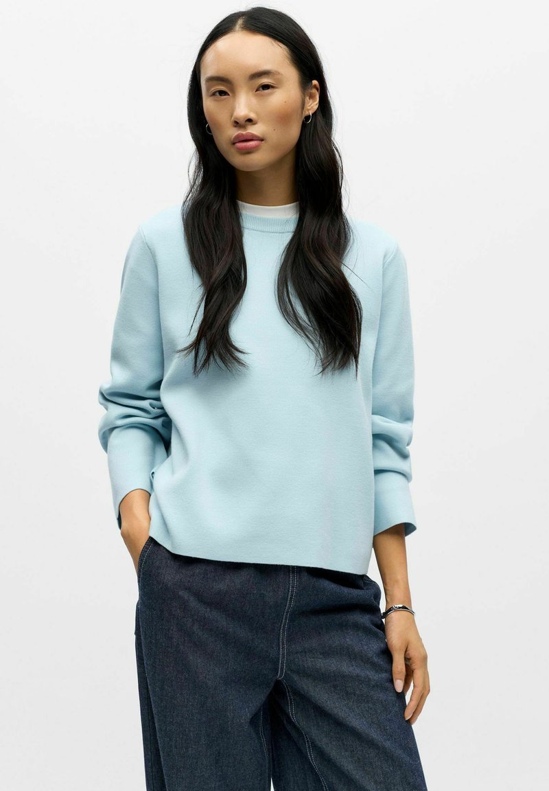 Jasnoniebieski sweatshirt z okrągłym dekoltem, długimi rękawami i luźnym krojem. W zestawieniu z ciemnymi dżinsami. Gładka struktura bez widocznych wzorów.