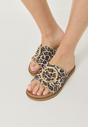 Pieds portant des sandales slides imprimé léopard avec de grandes boucles circulaires dorées sur des semelles couleur beige sur fond blanc.
